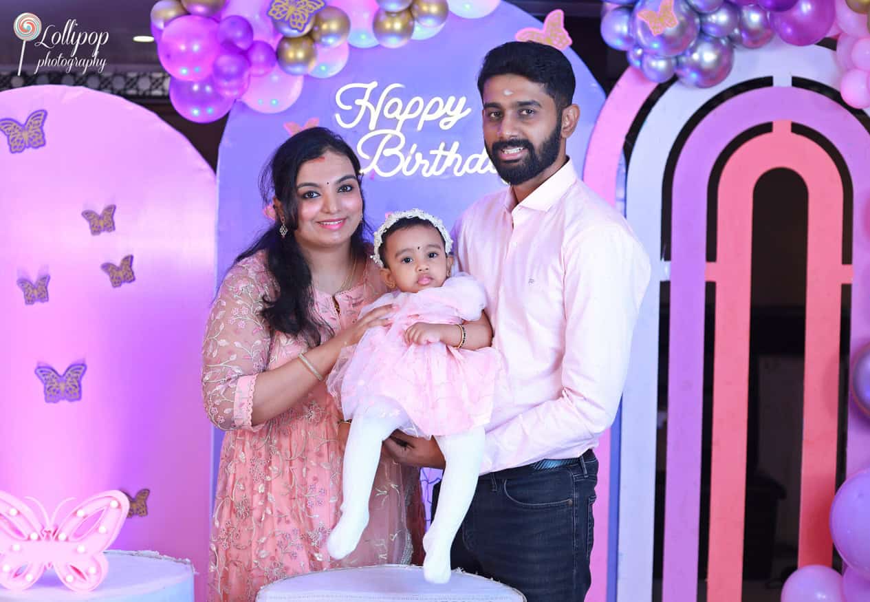 Smiling family of three posing in front of a colorful birthday setup with balloons and butterfly decorations, captured by Lollipop Photography in Chennai.