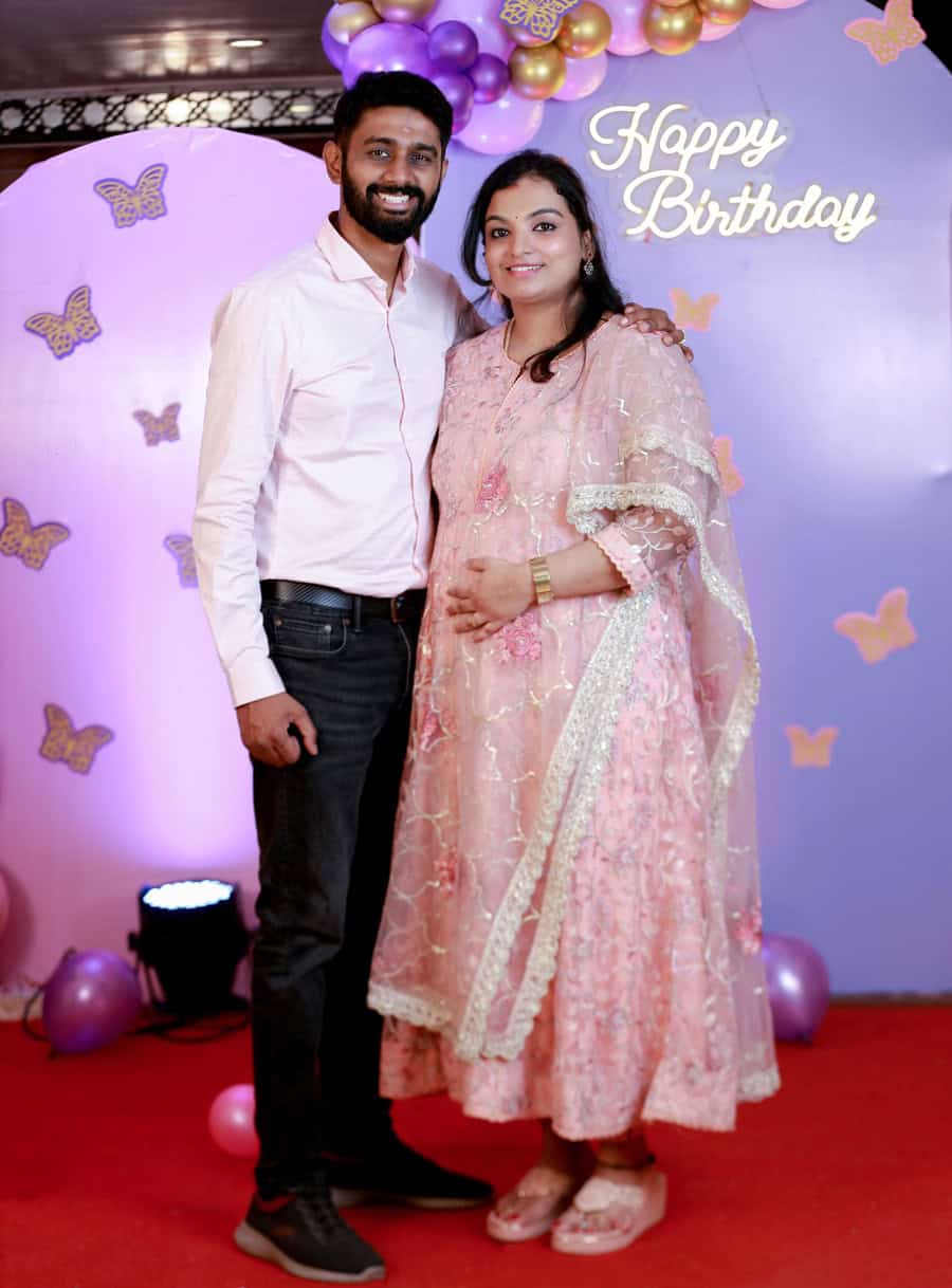 Smiling parents standing together with their baby girl at her birthday celebration, with a backdrop of festive balloons and butterflies in Chennai by Lollipop Photography.