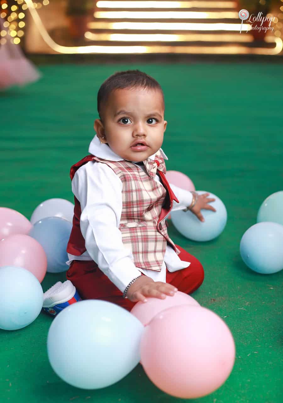 Adorable toddler dressed in a red vest and white shirt, surrounded by colorful balloons at a birthday event in Chennai, captured by Lollipop Photography