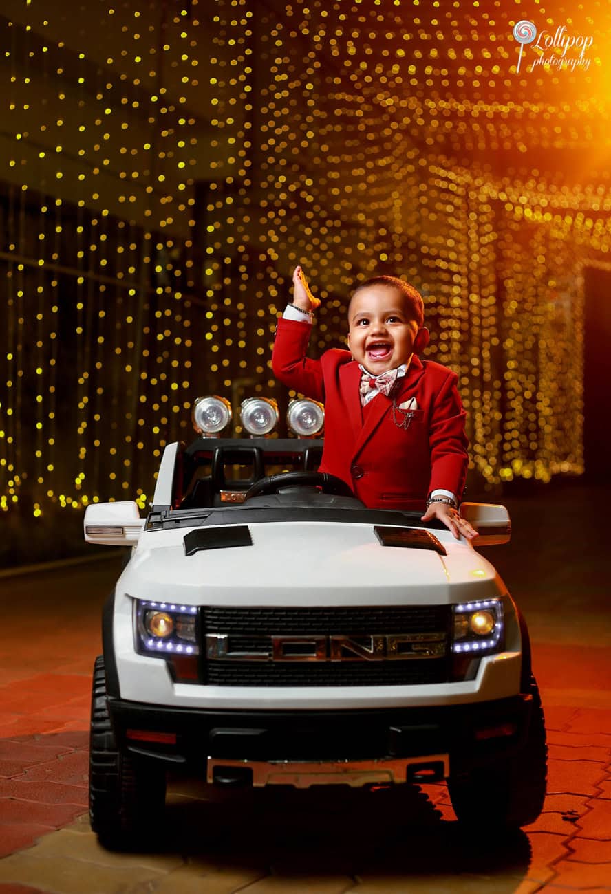 Joyous toddler in a red suit waves from his toy car, illuminated by a backdrop of sparkling lights at his birthday party, a magical night captured by Lollipop Photography in Chennai.