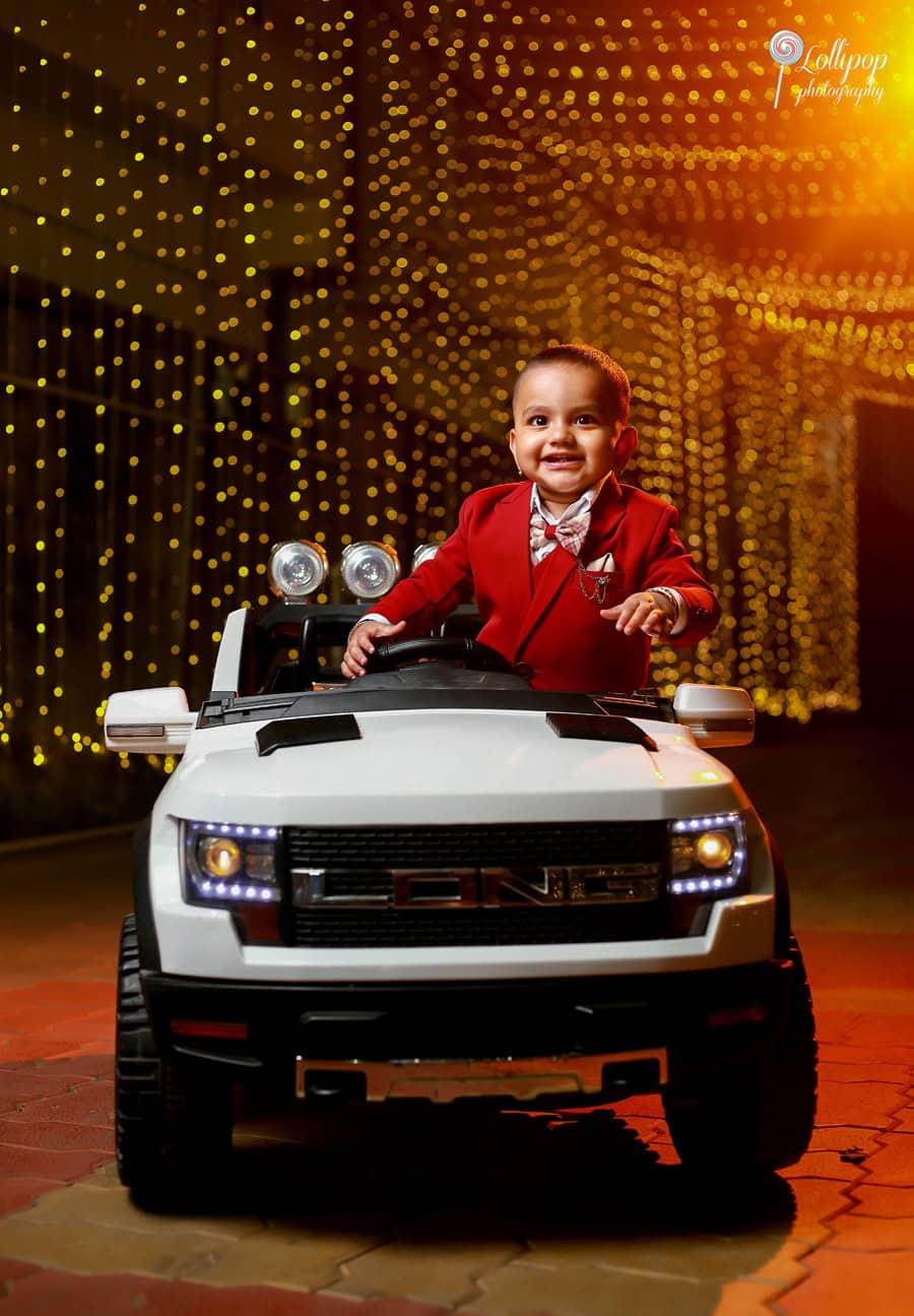 Cheerful toddler in a sharp red suit driving a toy car under a glittering light display at his birthday party, a charming moment preserved by Lollipop Photography in Chennai.