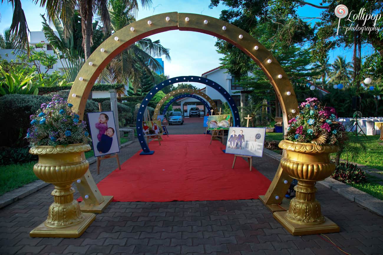 Elegant curved archway adorned with lights and family photos on display along a red carpet, welcoming guests to a birthday celebration in Chennai.