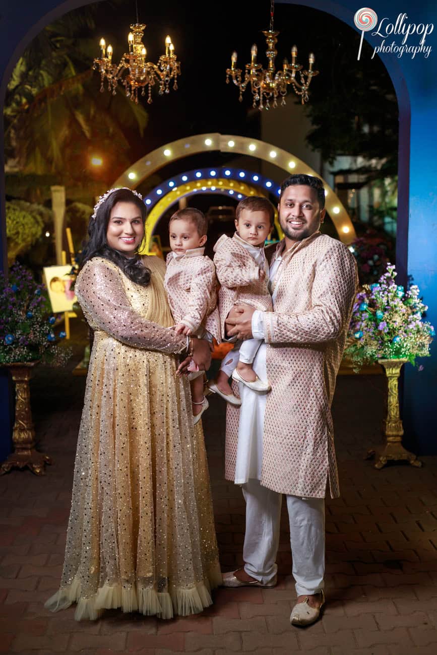 A family stands together in a festive outdoor setting under a chandelier, dressed in splendid traditional outfits, celebrating a birthday in Chennai.