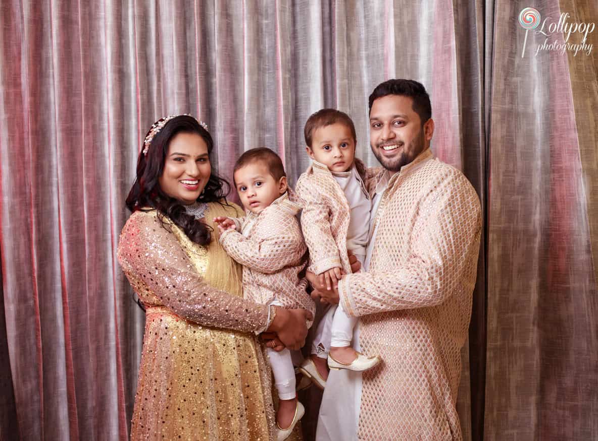 Family portrait in Chennai with a smiling mother and father holding their twin babies, all dressed in matching traditional Indian attire against a shimmering curtain backdrop.