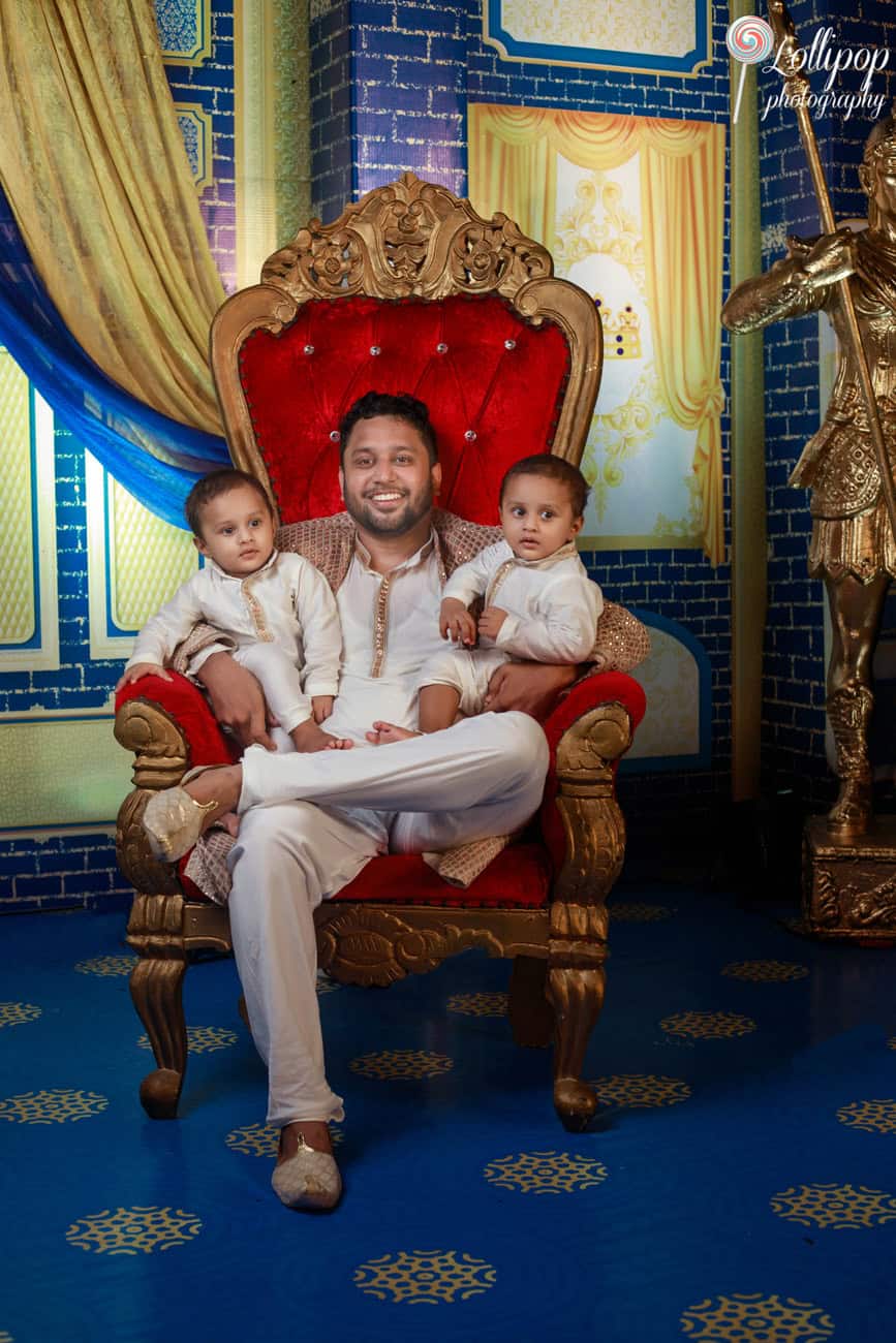 Father seated on a royal throne holding his twin boys during a birthday party in Chennai, all in white and gold traditional attire