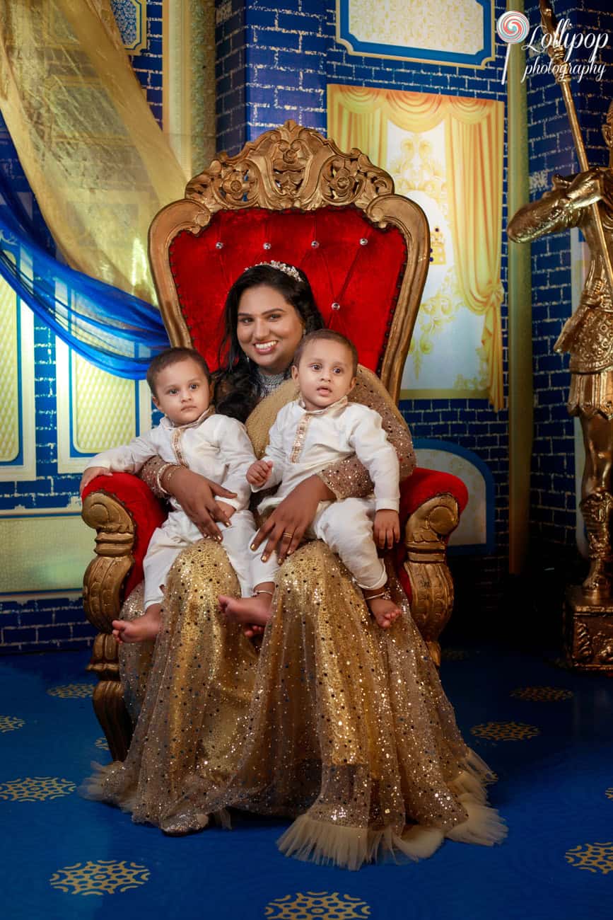 Mother seated on a red velvet throne at a birthday celebration, holding her twin boys dressed in traditional attire in Chennai.