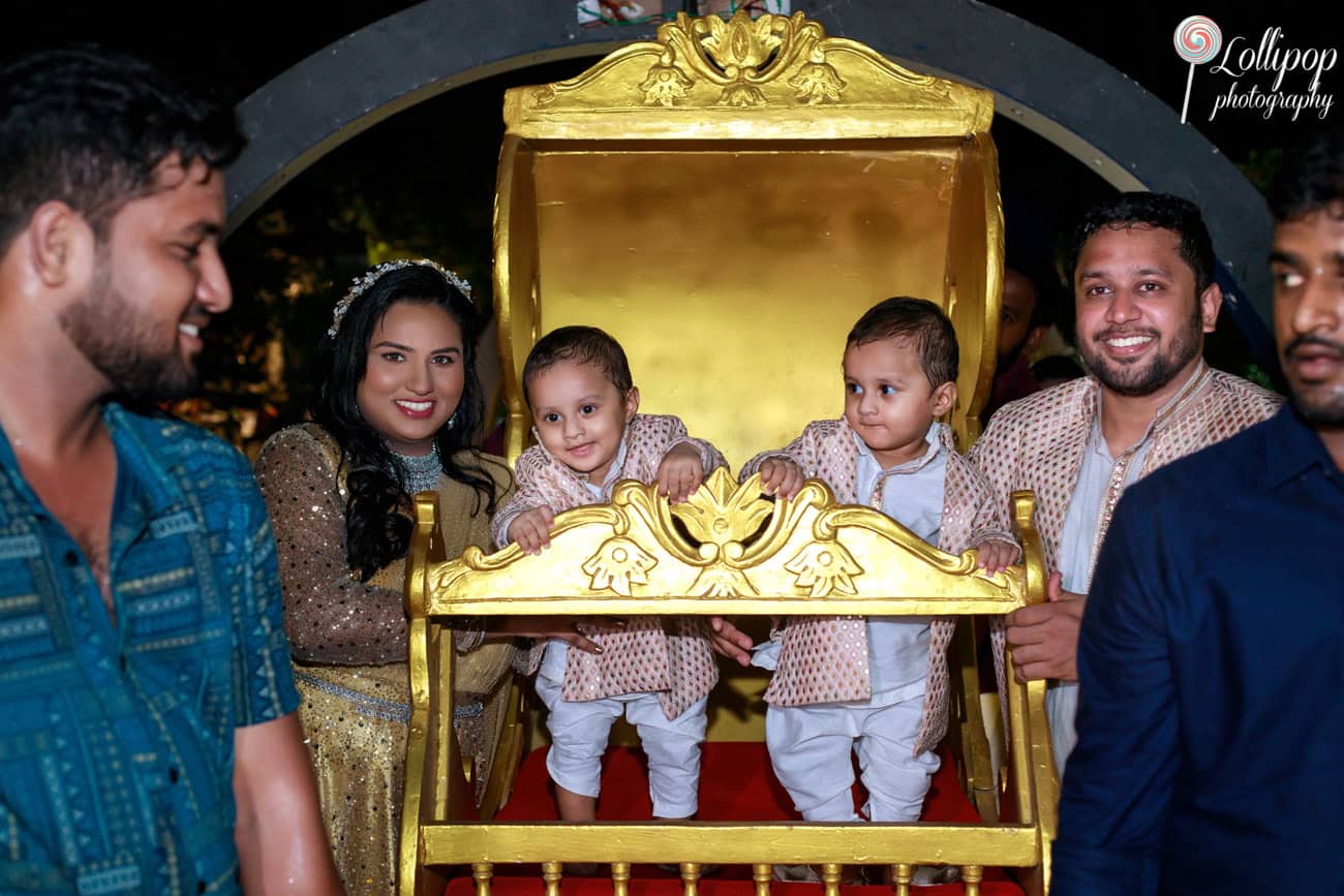 Twin toddlers enjoy a ride in a golden carriage during their birthday celebration, with parents smiling beside them, in Chennai.