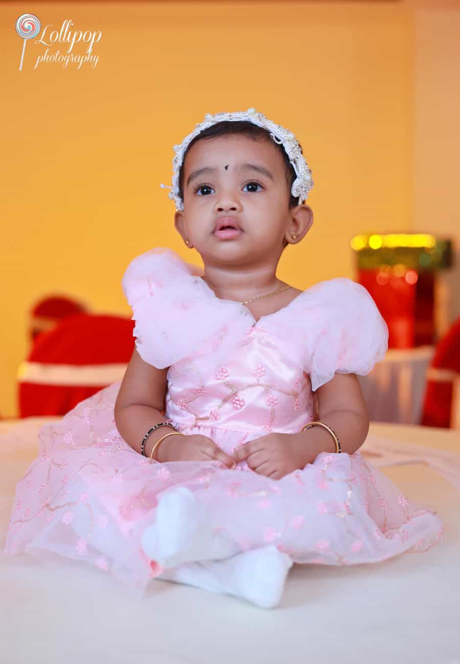 Baby girl dressed in a fluffy pink dress with flower embellishments, sitting calmly with a whimsical expression, photographed by Lollipop Photography.