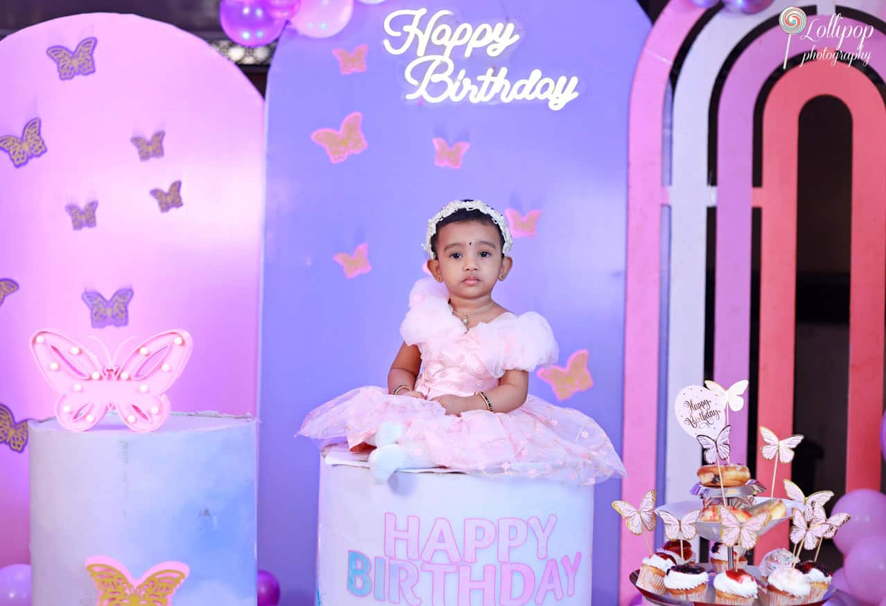 Baby girl celebrating her birthday seated on a large cake prop with a butterfly theme, surrounded by light pink and purple decorations, captured by Lollipop Photography.