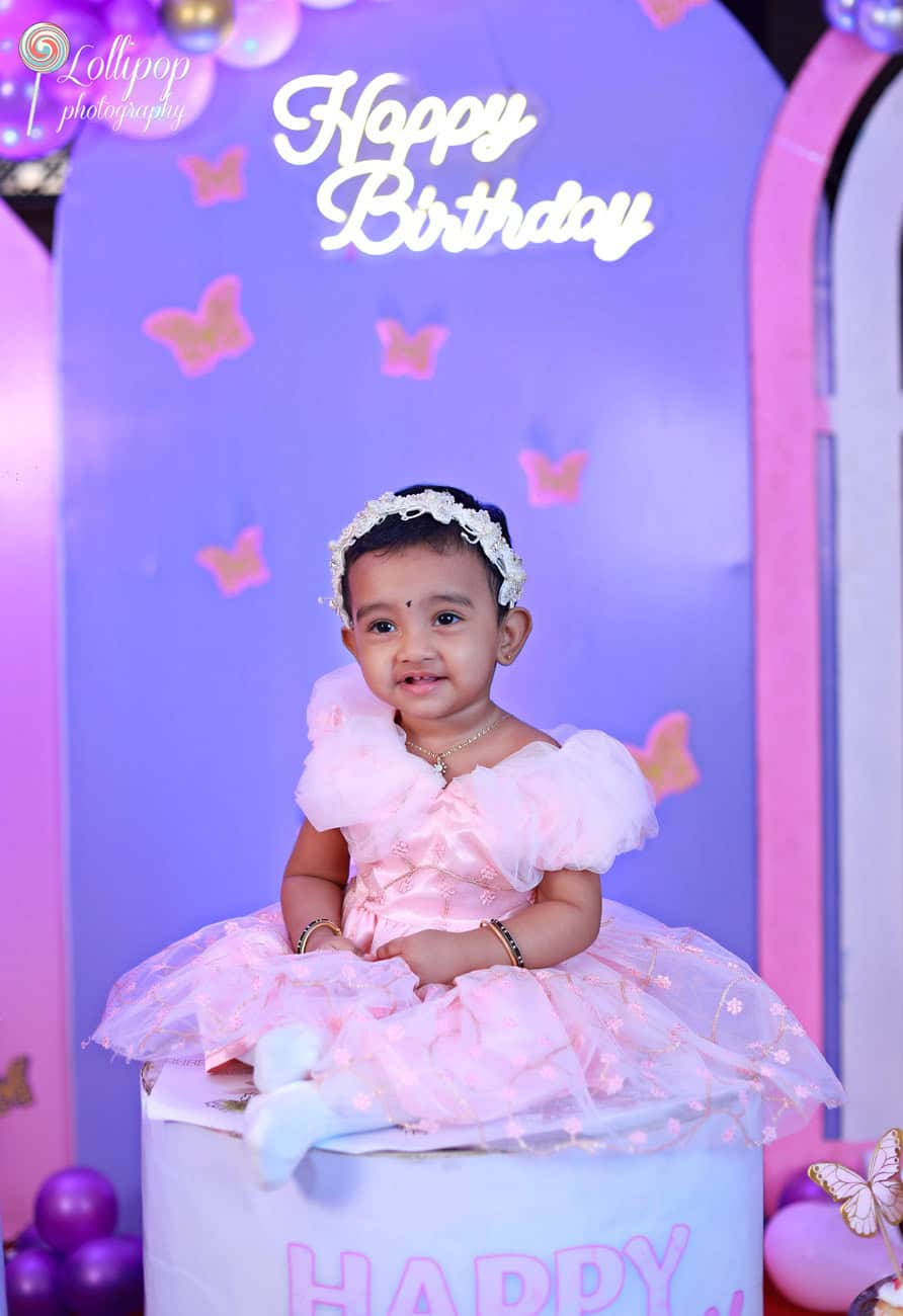 Baby girl in a beautiful pink dress smiling while seated on a birthday cake prop under a Happy Birthday sign, surrounded by butterfly decorations, by Lollipop Photography