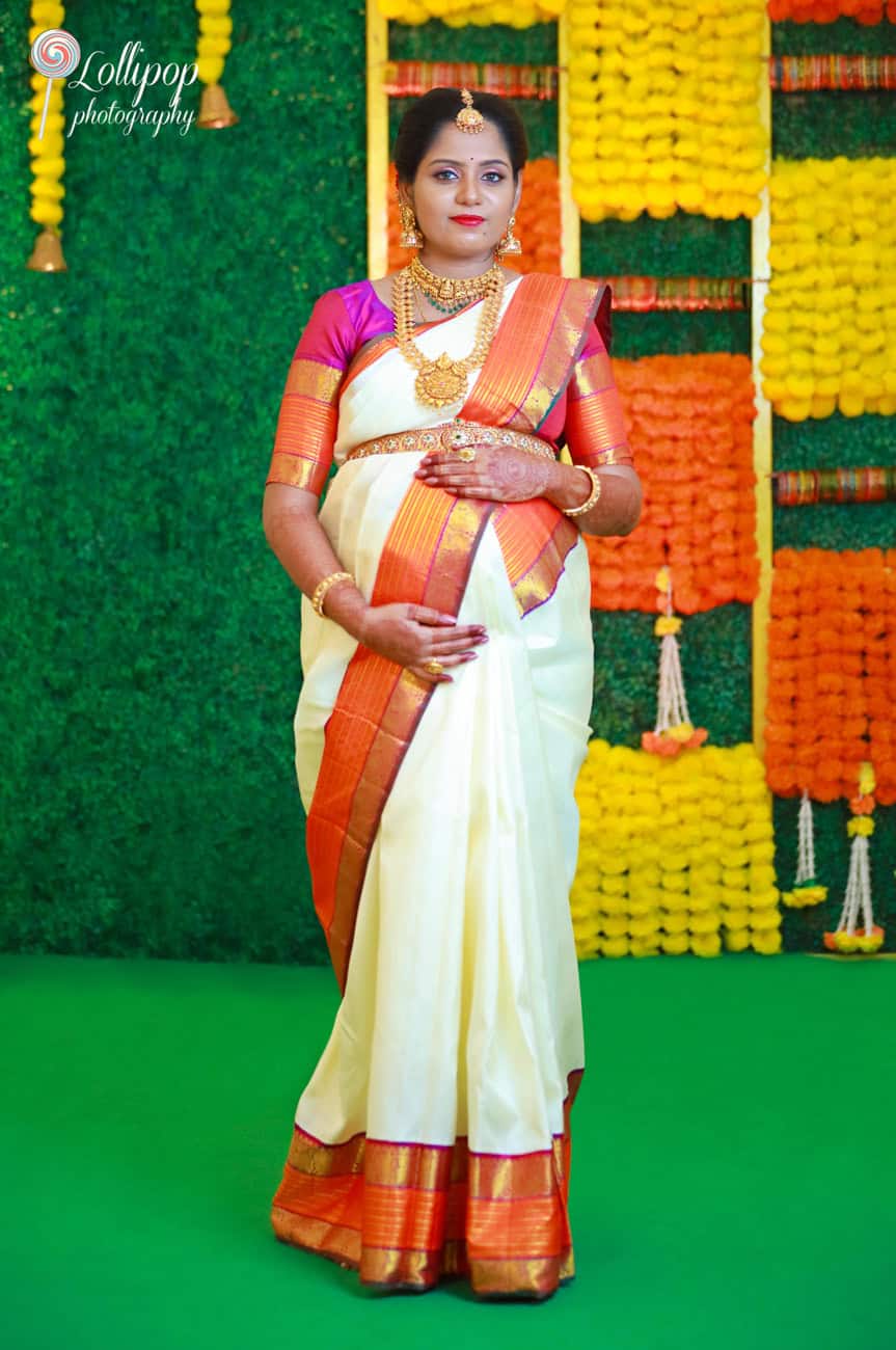 A radiant pregnant woman in a traditional saree stands confidently in front of a vibrant floral backdrop, capturing a moment of joy in Chennai.