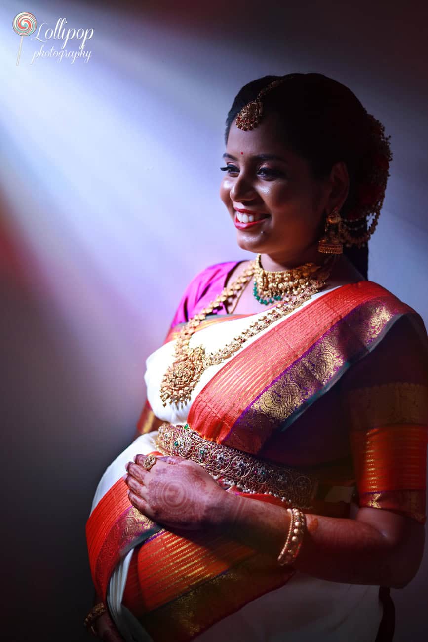 A joyful pregnant woman dressed in a vibrant saree smiles broadly, bathed in soft lighting in Chennai.