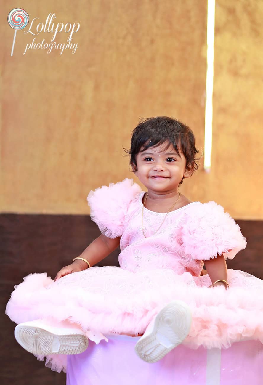 Little Kanali, dressed in a soft pink dress, smiles joyfully as she celebrates her first birthday, beautifully captured by Lollipop Photography in Chennai.