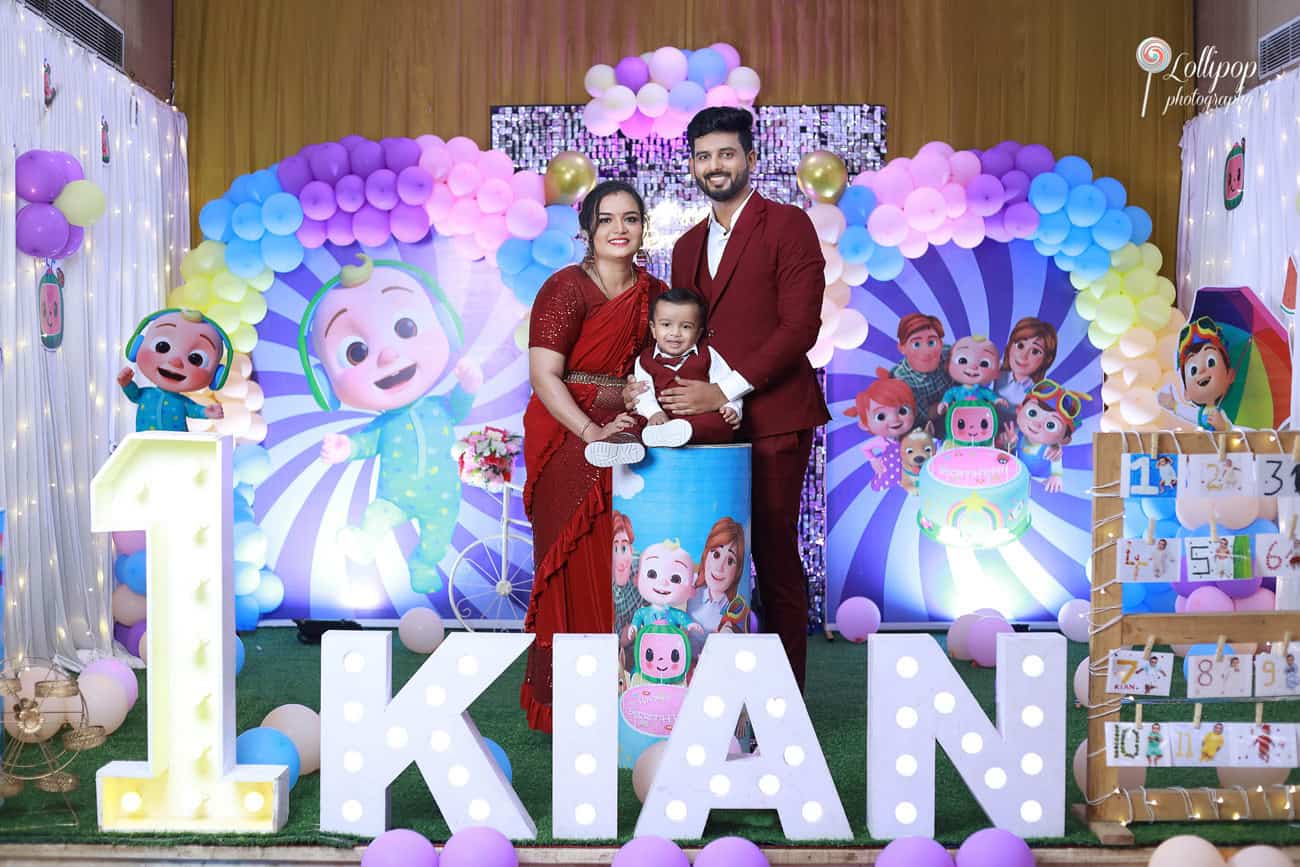 Kian and family in front of a colorful character-themed backdrop, with '1' and 'Kian' light-up letters, celebrating his first birthday