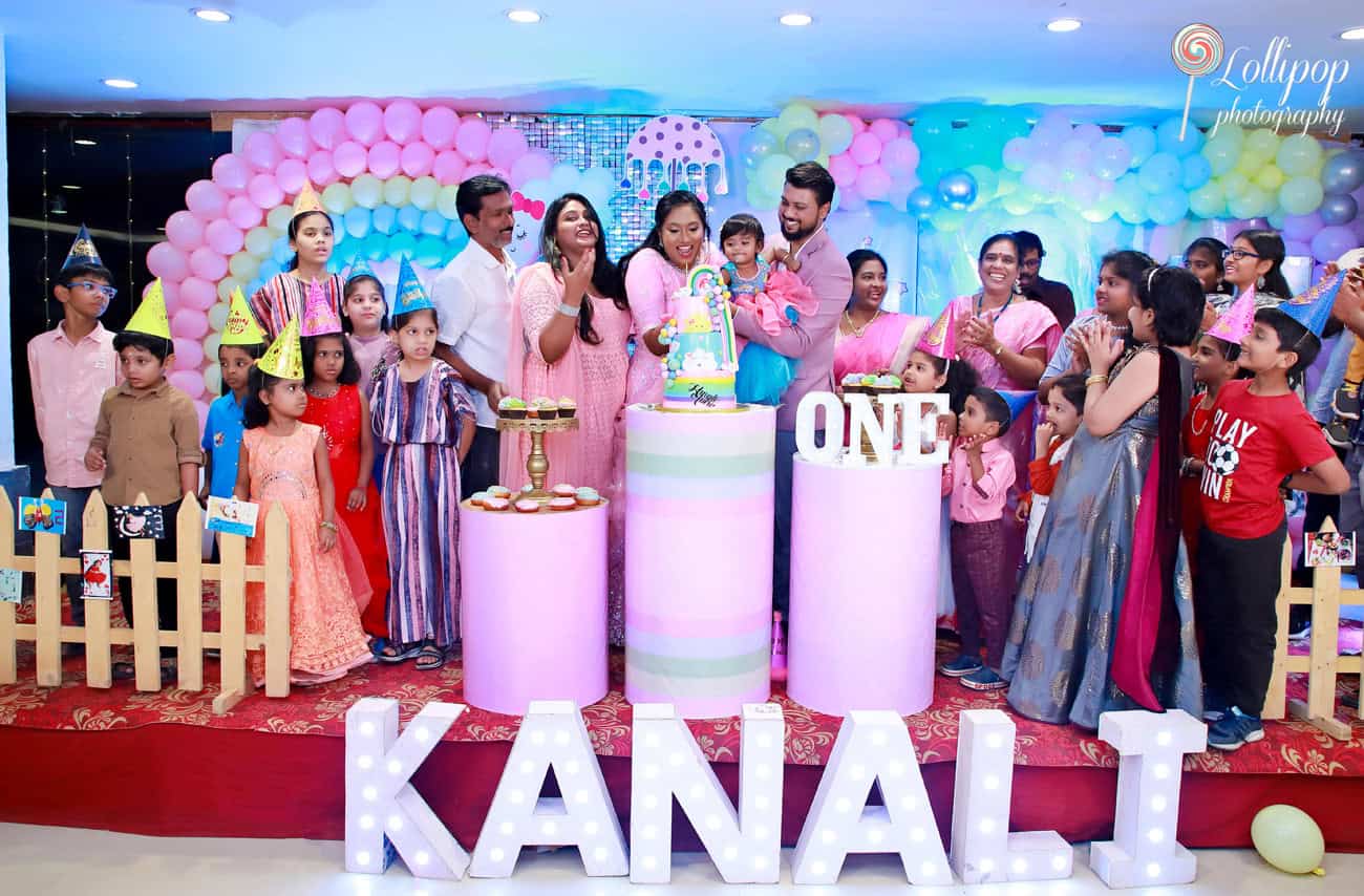 Kanali’s family celebrating her birthday around a rainbow cake, with children wearing party hats, captured by Lollipop Photography