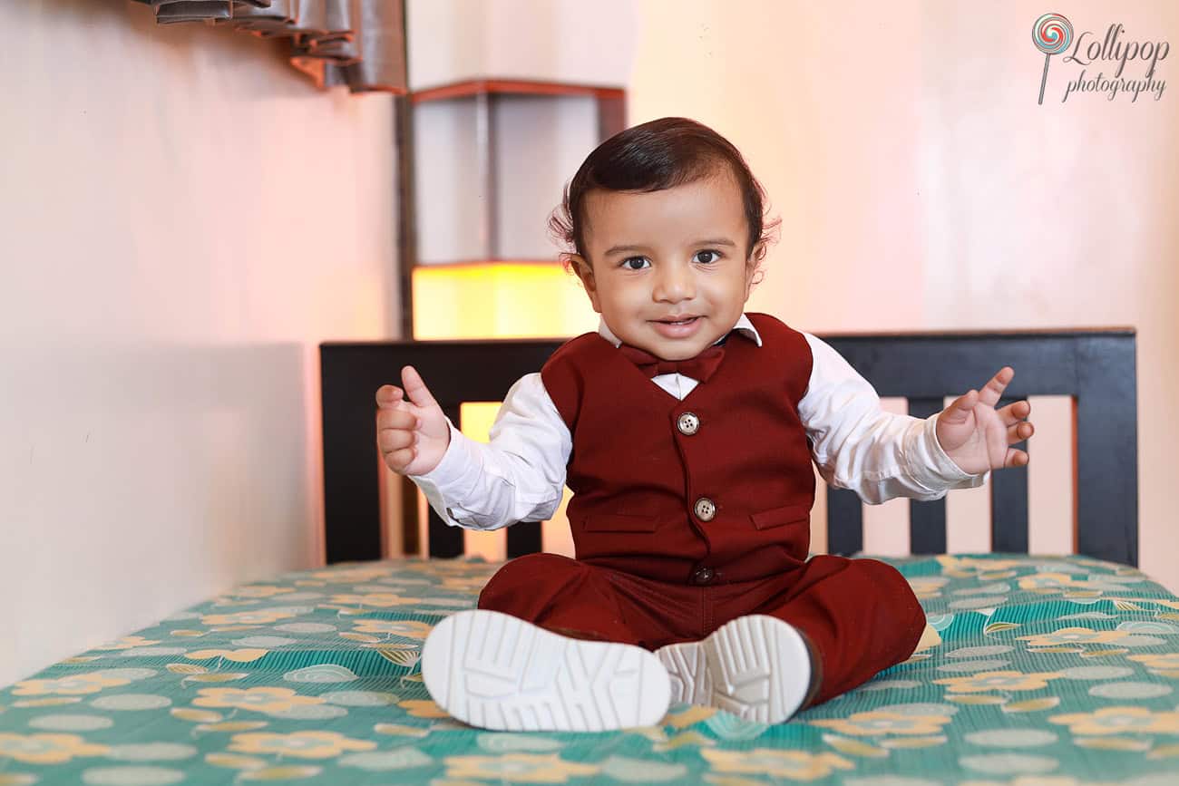 Adorable Kian seated on a bed, dressed in a maroon suit with a white shirt, spreading joy during his first birthday photoshoot in Chennai.