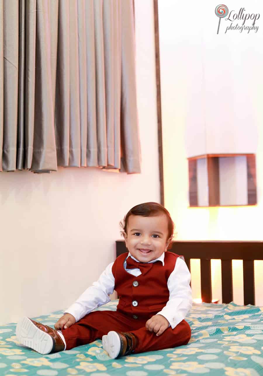 Kian dressed in a maroon suit, smiling brightly during his first birthday photoshoot in Chennai, sitting on a patterned bed and creating a cheerful moment captured by Lollipop Photography