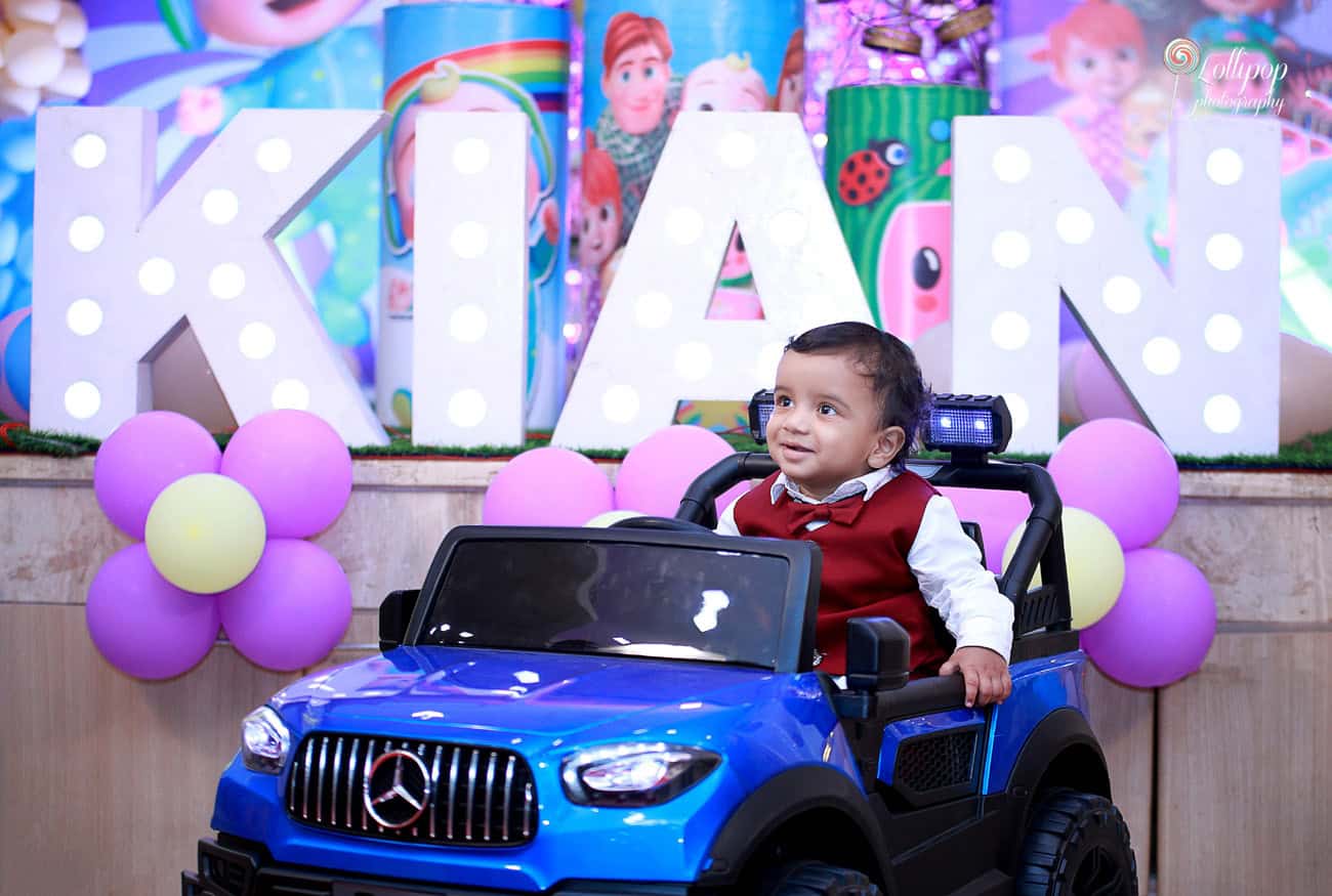 Kian sits in a blue toy car with a charming smile during his birthday celebration in Chennai, surrounded by balloons and decor by Lollipop Photography.