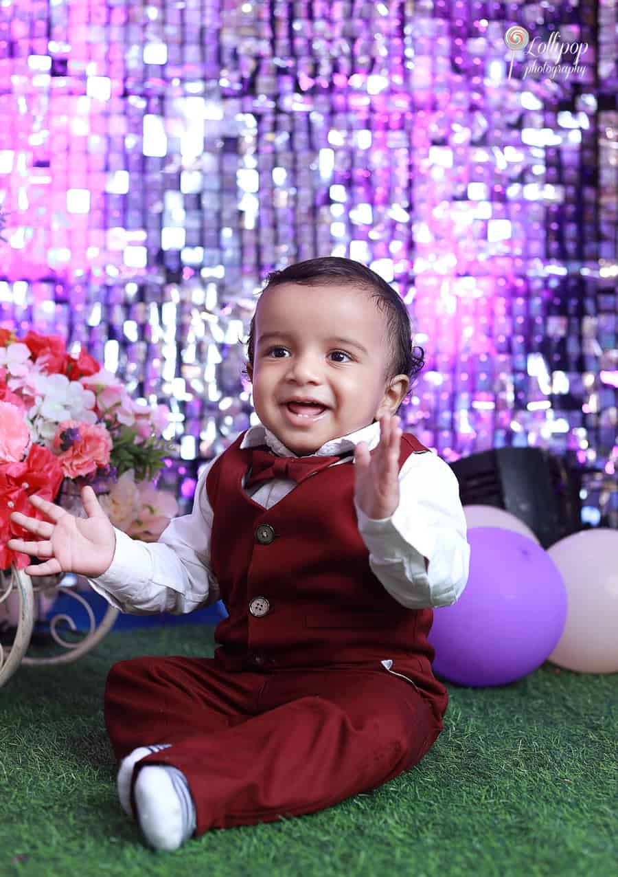 Kian spreading smiles while celebrating his first birthday, with a shimmering purple backdrop, photographed by Lollipop Photography in Chennai