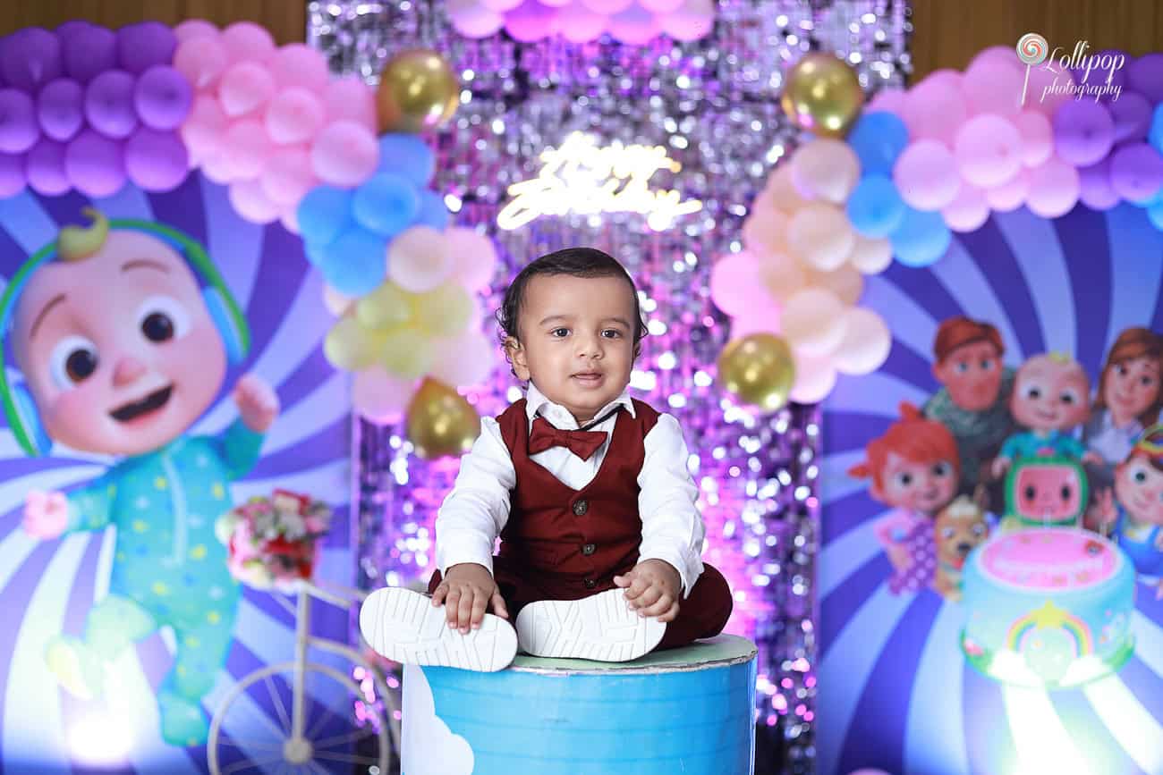 Kian sitting adorably on a blue barrel in front of a vibrant birthday-themed backdrop, captured by Lollipop Photography during his first birthday celebration in Chennai.