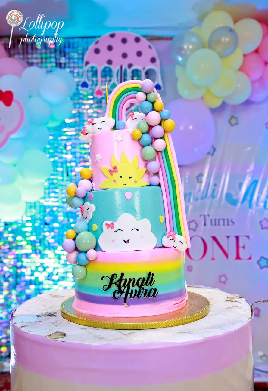 Vibrant rainbow-themed birthday cake featuring smiling clouds and a sun, created for Kanali's first birthday by Lollipop Photography