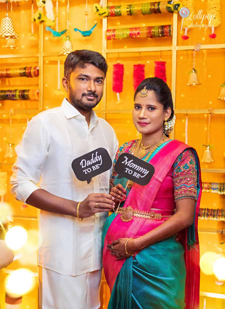 Arthi and Naveen Kumar celebrating their baby shower in Chennai, holding signs that say 'Daddy to be' and 'Mommy to be,' captured by Lollipop Photography.