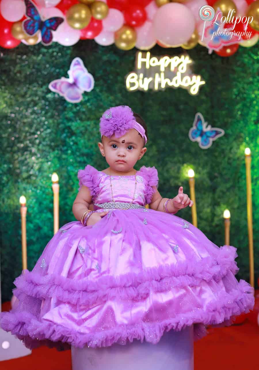 An adorable baby girl in a purple dress standing at her birthday celebration stage with butterfly decor and balloons. Birthday photoshoot by Lollipop Photography, Chennai