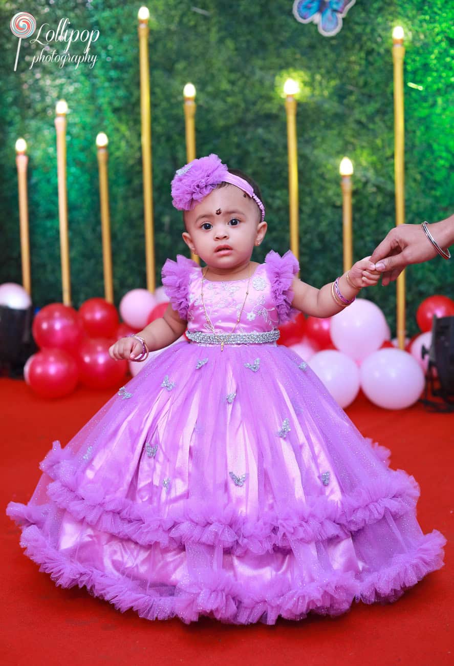 A baby girl dressed in a beautiful purple gown stands on the red carpet of a butterfly-themed birthday setup. Captured by Lollipop Photography, Chennai.