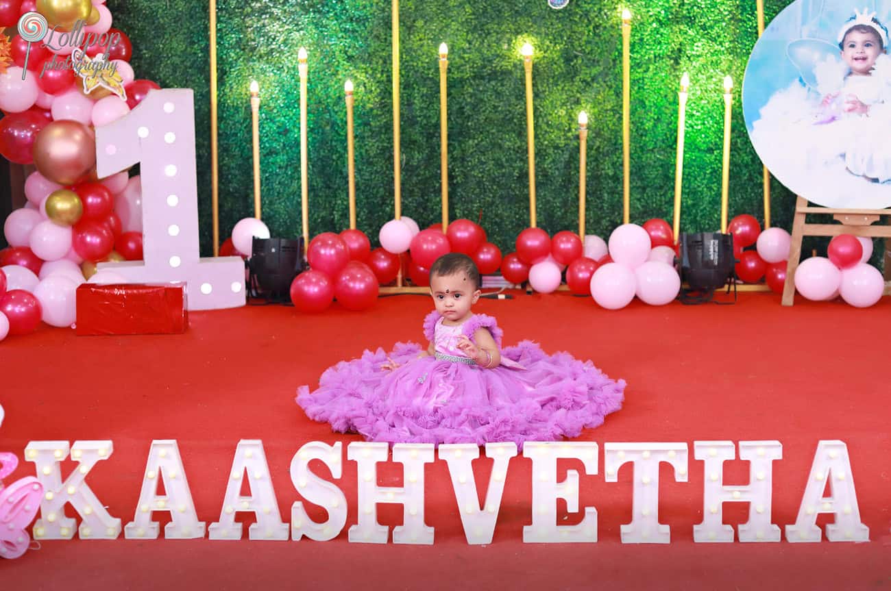 Baby girl in a stunning purple gown posing in front of a custom birthday stage with her name illuminated. Birthday photoshoot by Lollipop Photography, Chennai.