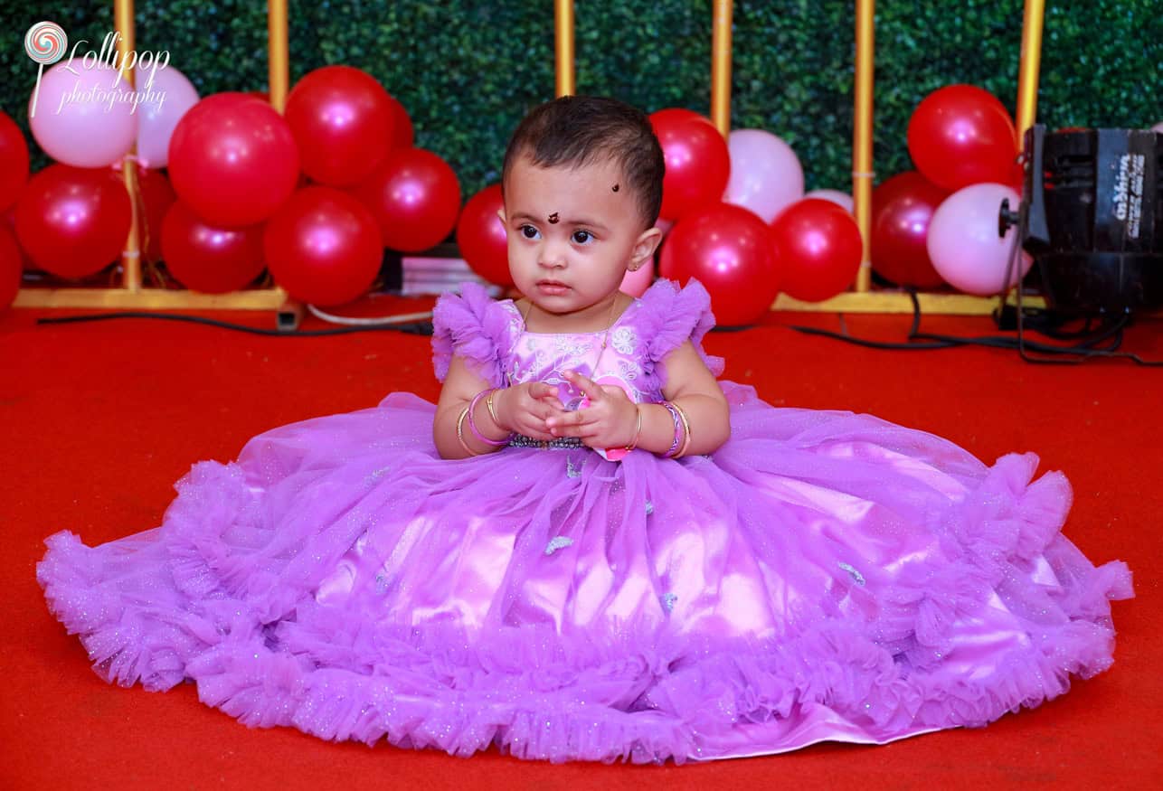 A baby girl in a purple gown sitting on the red carpet surrounded by balloons at her butterfly-themed birthday. Captured by Lollipop Photography, Chennai