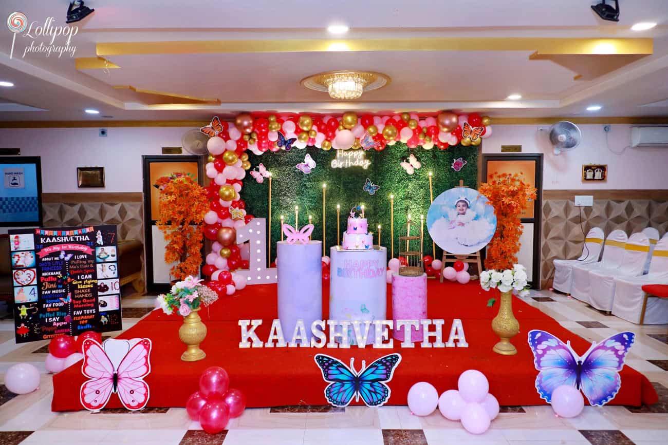 A beautifully decorated birthday stage with a butterfly theme, featuring vibrant balloons, a custom name display, and a tiered cake. Captured by Lollipop Photography in Chennai