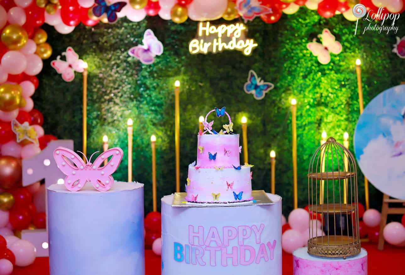 Close-up of a pink butterfly-themed birthday cake on a vibrant stage, surrounded by candle lights and balloons. Lollipop Photography birthday shoot in Chennai