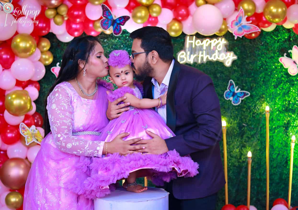 A heartwarming family moment with parents kissing their baby girl during her butterfly-themed birthday celebration. Captured by Lollipop Photography, Chennai."