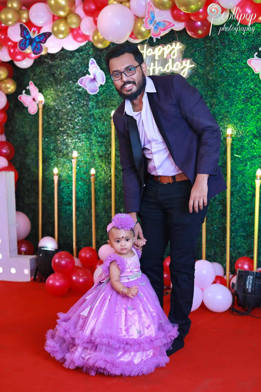 A proud father posing with his baby girl in a purple gown during her butterfly-themed birthday celebration. Captured by Lollipop Photography, Chennai