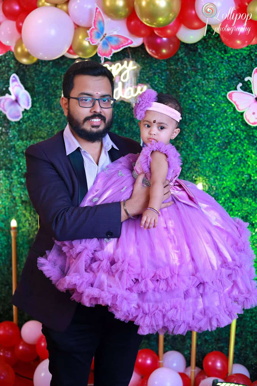 Father holding his baby girl dressed in a purple gown with butterfly decor in the background at her birthday celebration. Birthday photoshoot by Lollipop Photography, Chennai