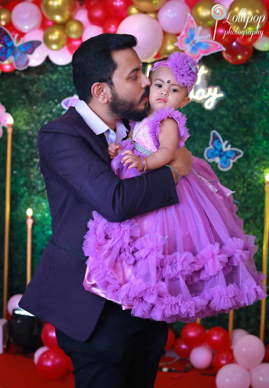 A father kissing his baby girl in a lavender gown amidst a colorful birthday backdrop. Captured by Lollipop Photography, Chennai