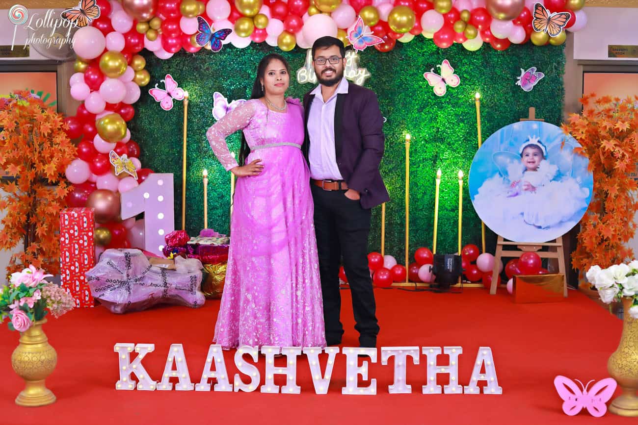 Parents posing in front of a vibrant butterfly-themed birthday stage with colorful decor. Captured by Lollipop Photography, Chennai.