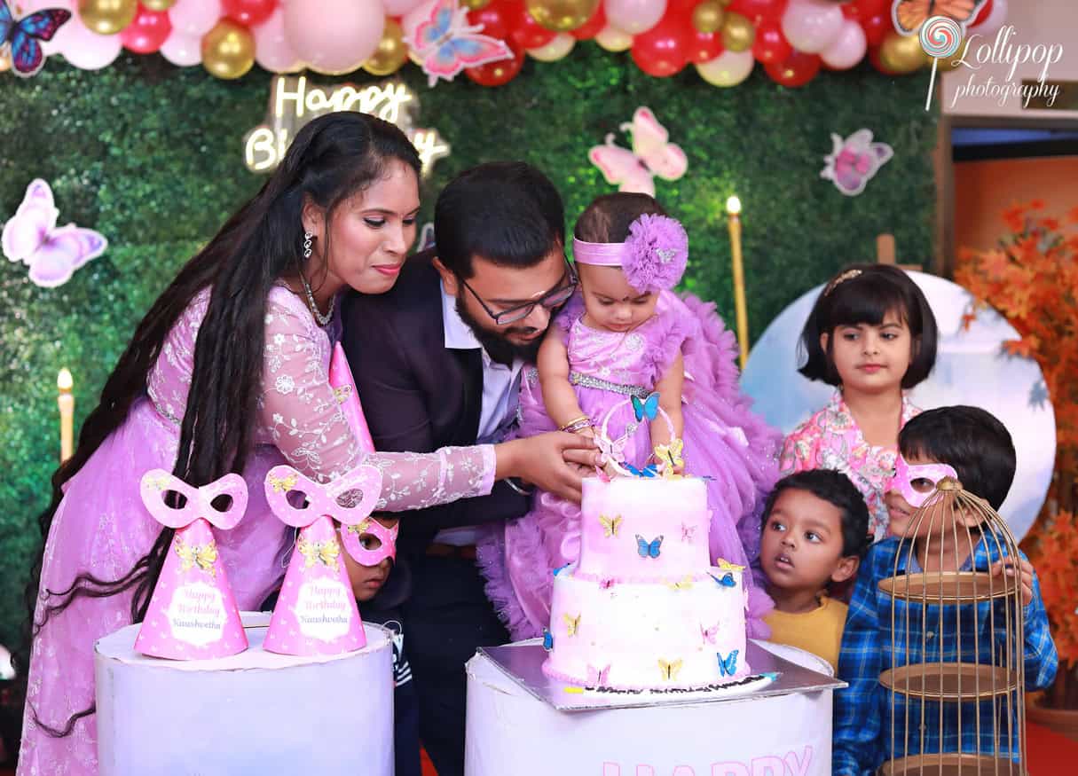 Parents helping their baby girl cut her butterfly-themed birthday cake surrounded by family and friends. Birthday photoshoot by Lollipop Photography, Chennai.