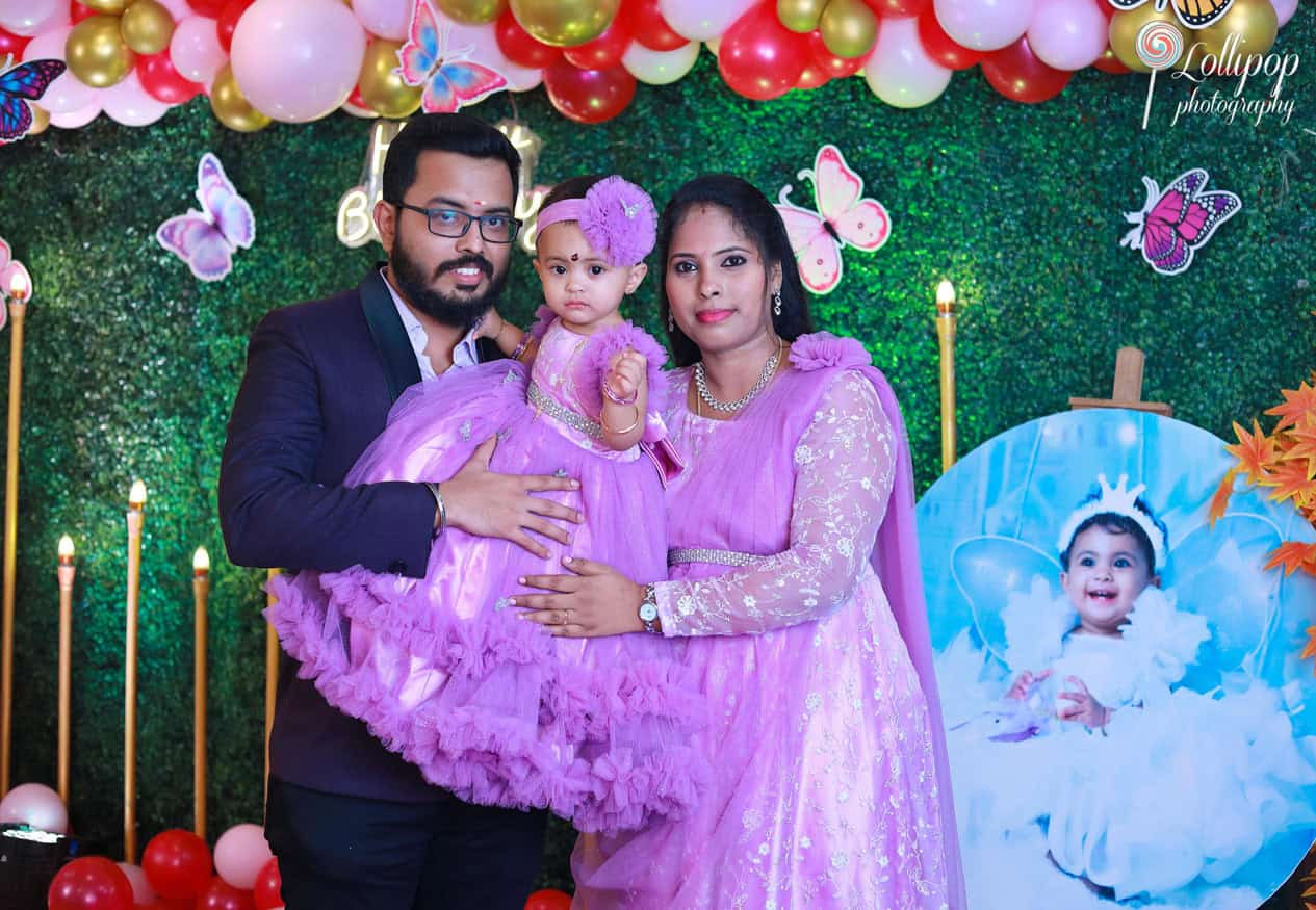 Parents holding their baby girl in front of a colorful butterfly-themed backdrop during her birthday celebration. Captured by Lollipop Photography, Chennai.