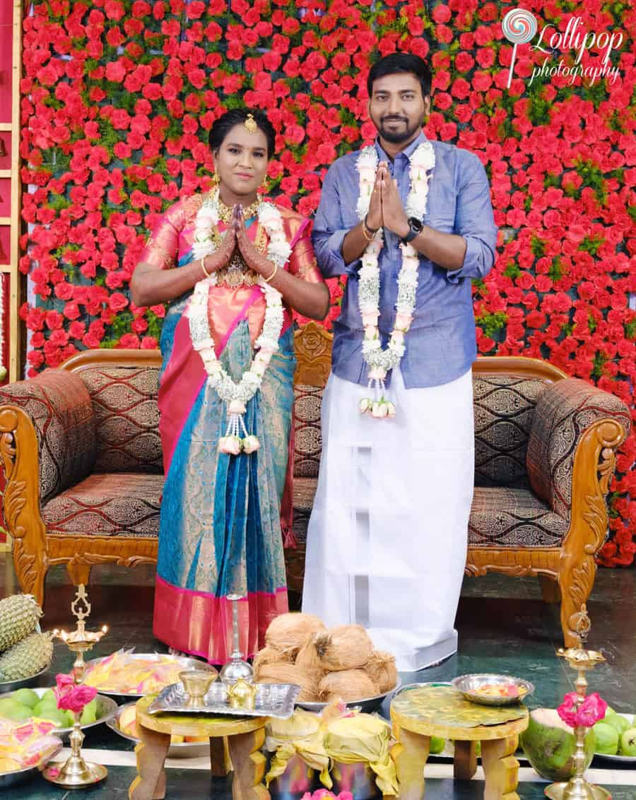 Aswini and her spouse pose in traditional attire, hands folded in a respectful gesture, against a vibrant red floral backdrop at their baby shower in Chennai.