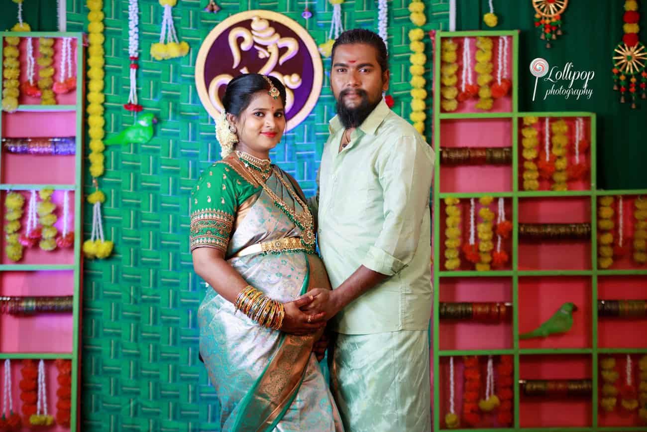 Sharmila and her husband share a sweet moment holding hands on the baby shower stage, surrounded by vibrant decor, captured by Lollipop Photography in Coimbatore.