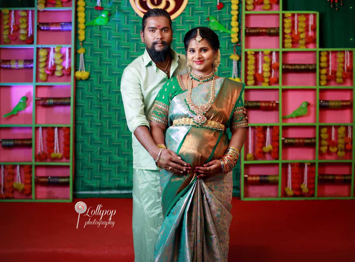 Sharmila and her husband pose gracefully in traditional attire, with vibrant decor in the background, celebrating her baby shower in Coimbatore, by Lollipop Photography.