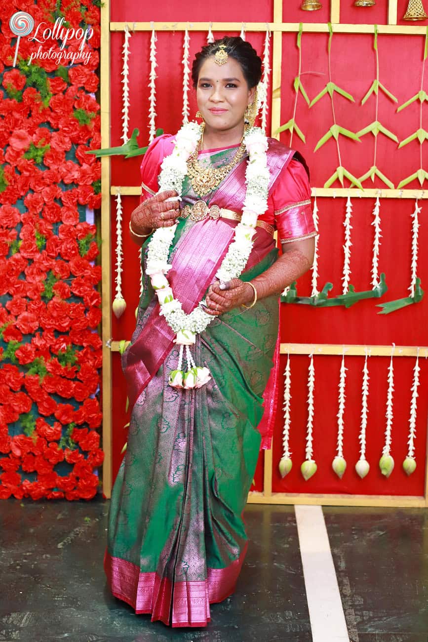 A soon-to-be mother in a pink saree adorned with traditional garlands poses gracefully against a backdrop of vibrant flowers, captured by Lollipop Photography in Chennai.